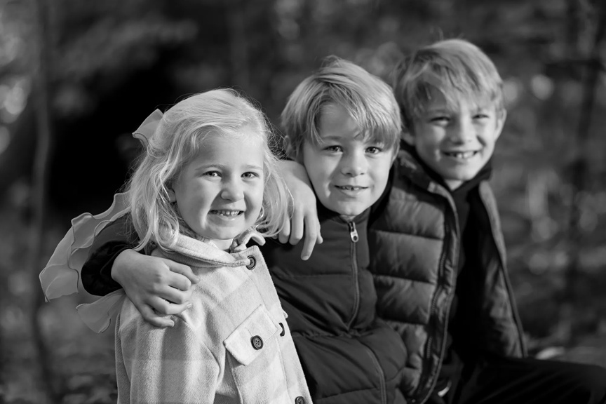 One girl and two boys are outside, arms wrapped around each other's shoulders. They are smiling and wearing coats.