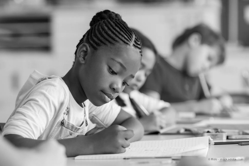 An elementary aged girl sits at her classroom desk, writing  on a notebook. A boy student is in the background behind her and is also writing in a notebook.