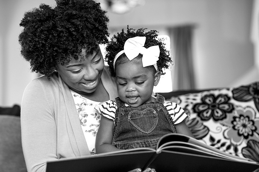 A mom is smiling, reading to the toddler seated on her lap.