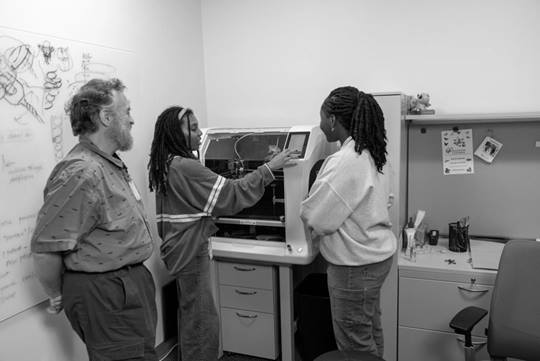A smiling employee leans against the wall and observes as two female interns learn how to use the 3D printer.