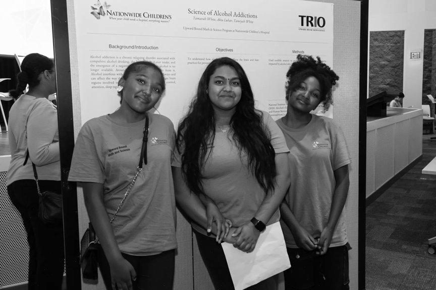 Three smiling female students stand in front of a large poster