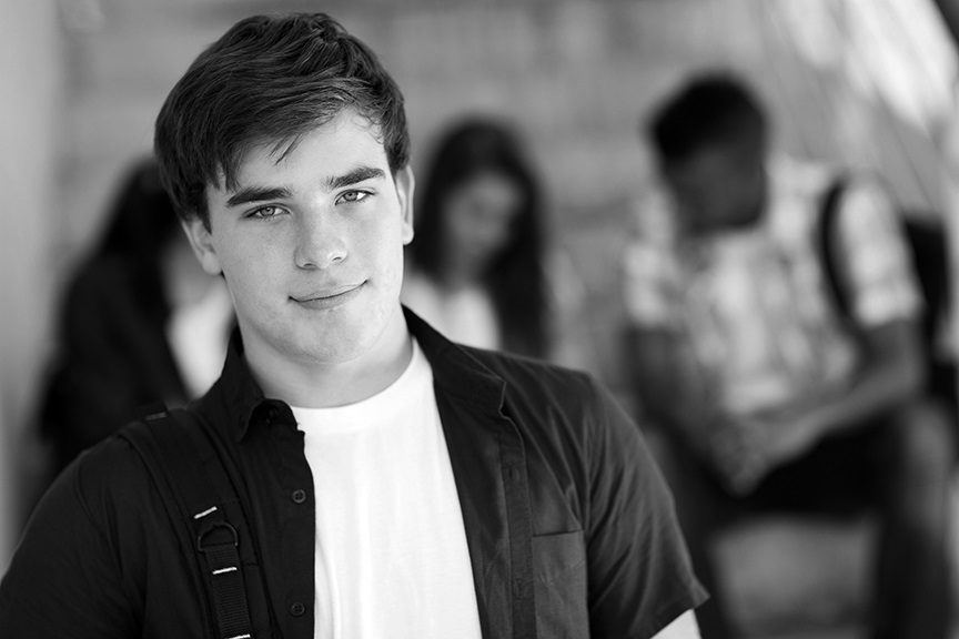 Adolescent standing in a hallway at school with friends chatting in the background