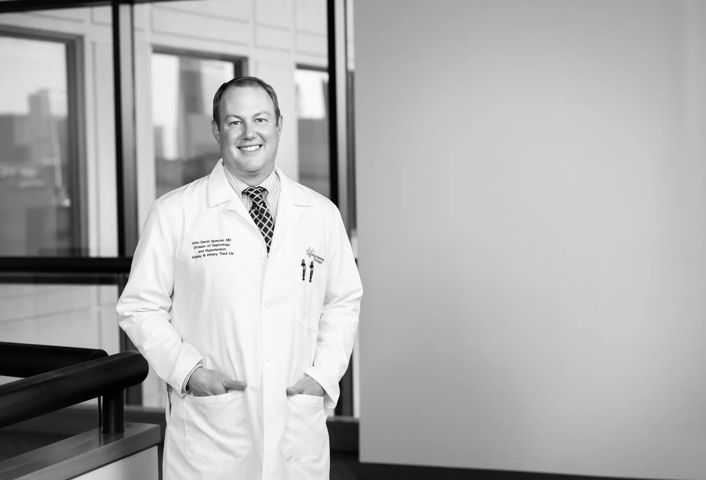 John Spencer is wearing a white coat with a shirt and tie underneath. He has his hands in his coat pockets and is smiling. Windows are shown in the hallway behind him.