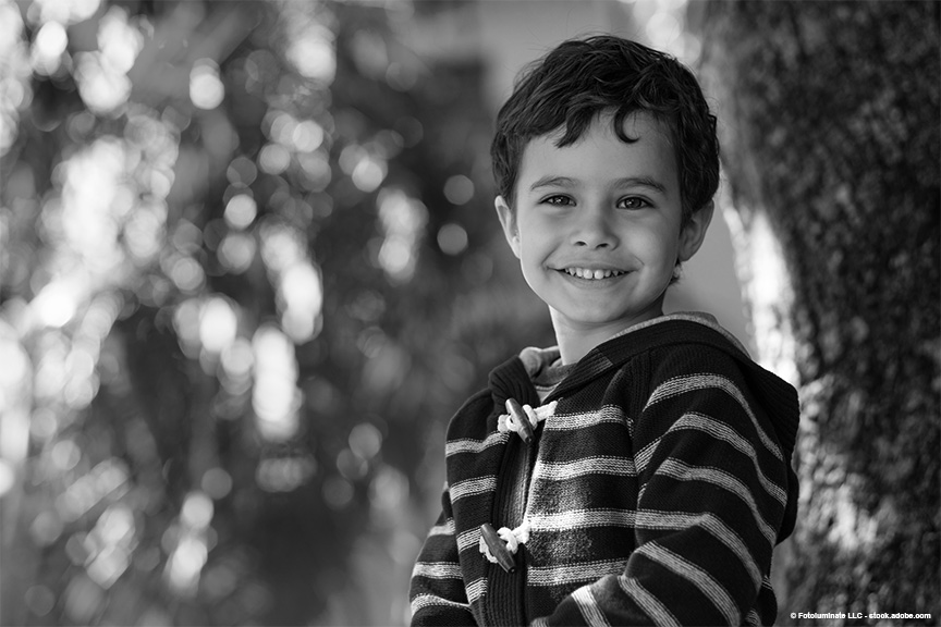 A smiling elementary aged boy is siting by a tree. He's wearing a striped zip up sweater.