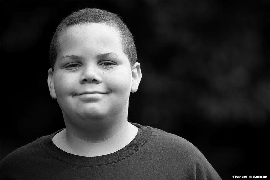 Older elementary-aged boy is smiling softly at the camera with a dark, solid background behind him.