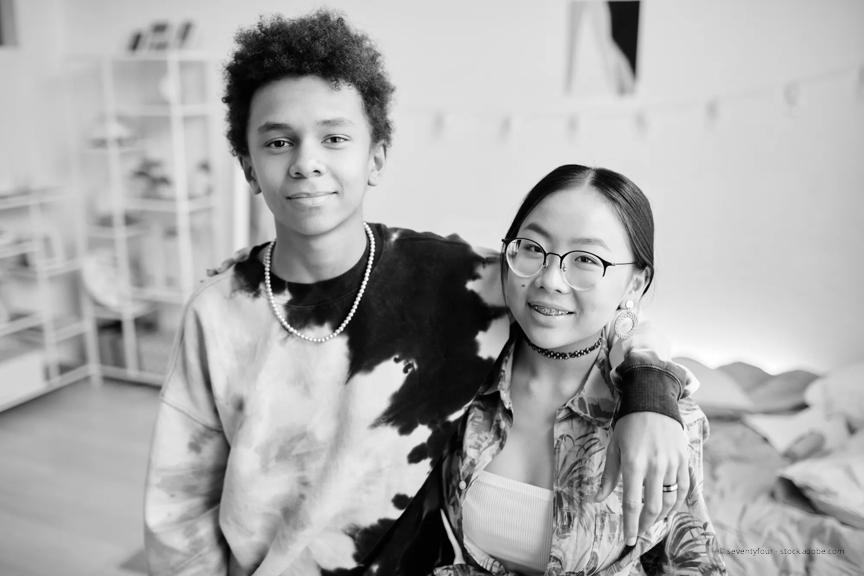 A teen boy wears a tie dye shirt and a chain around his neck. His left arm is resting on a teen girl's shoulder. She has glasses and braces. Both are smiling. 