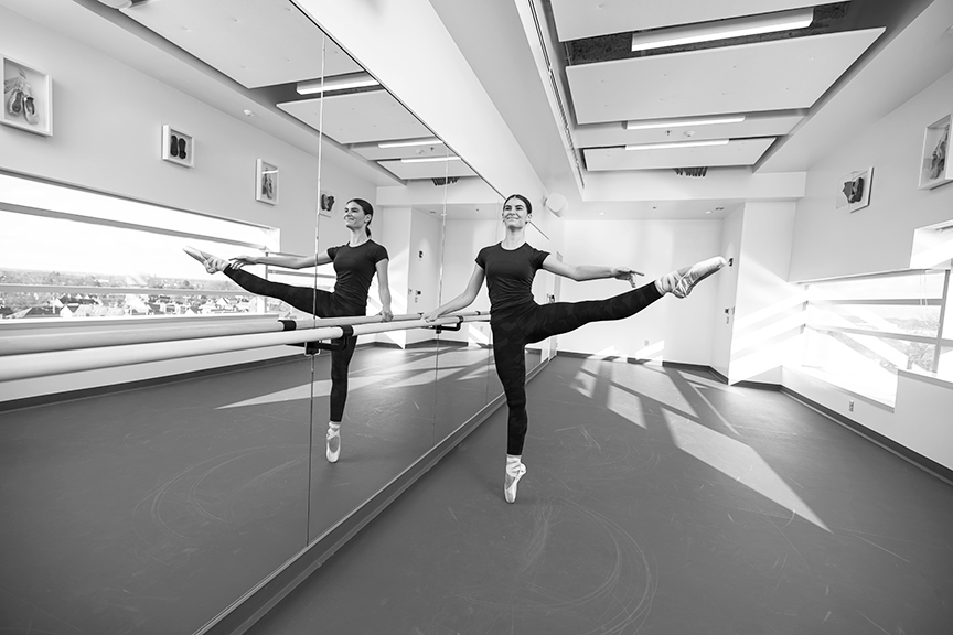A dancer wears a dark leotard and tights. She's en pointe, at the barre, in a studio setting.