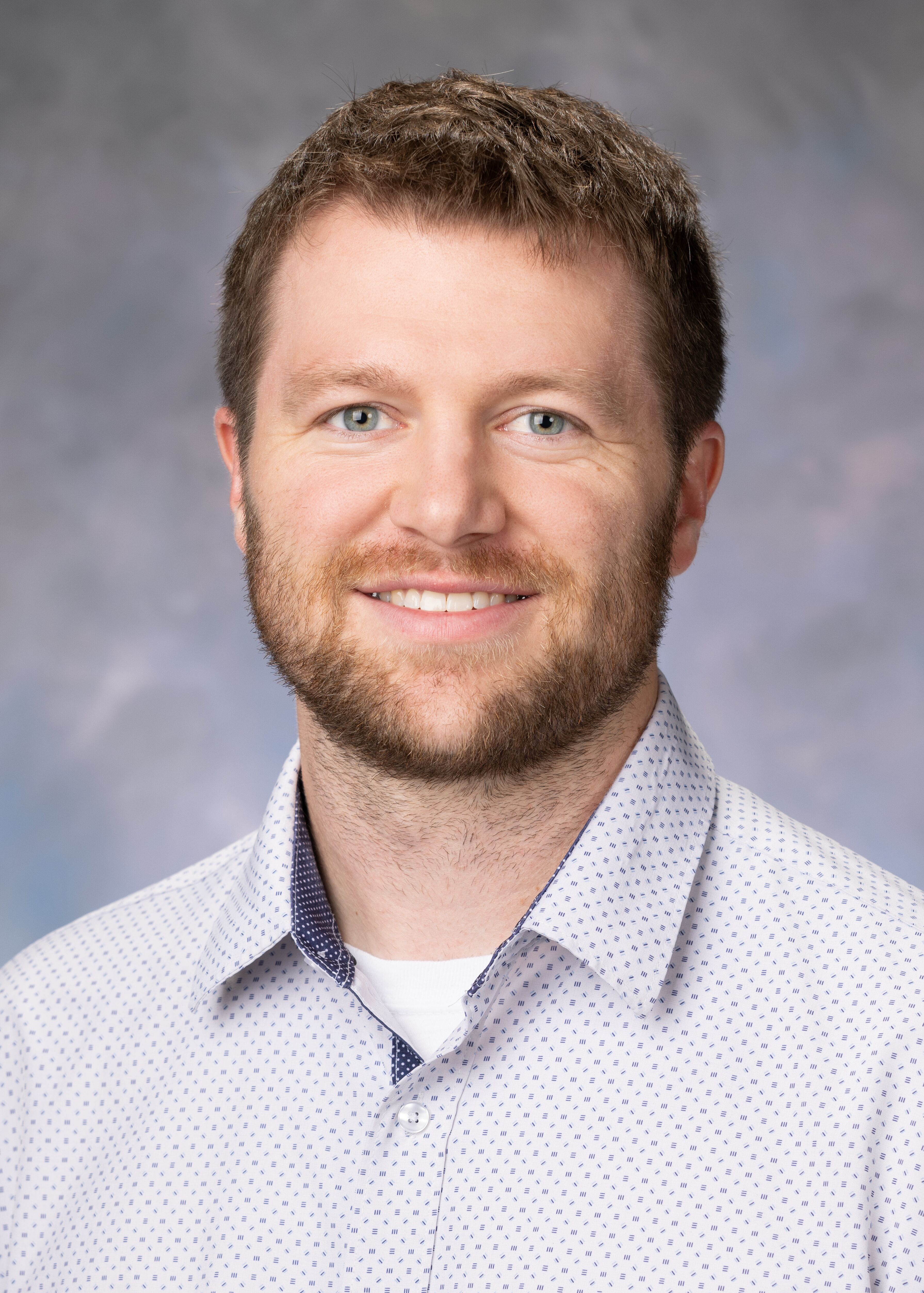 Portrait of Tyler Walsh. He had brown hair and a beard. He is wearing a light gray button up shirt with a white undershirt.