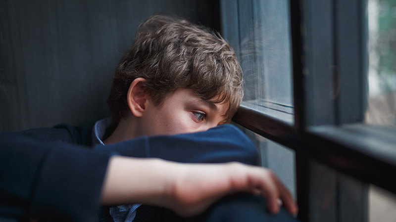 Young boy with curly hair sits quietly by a window, resting his head on his arm and gazing outside with a thoughtful expression.